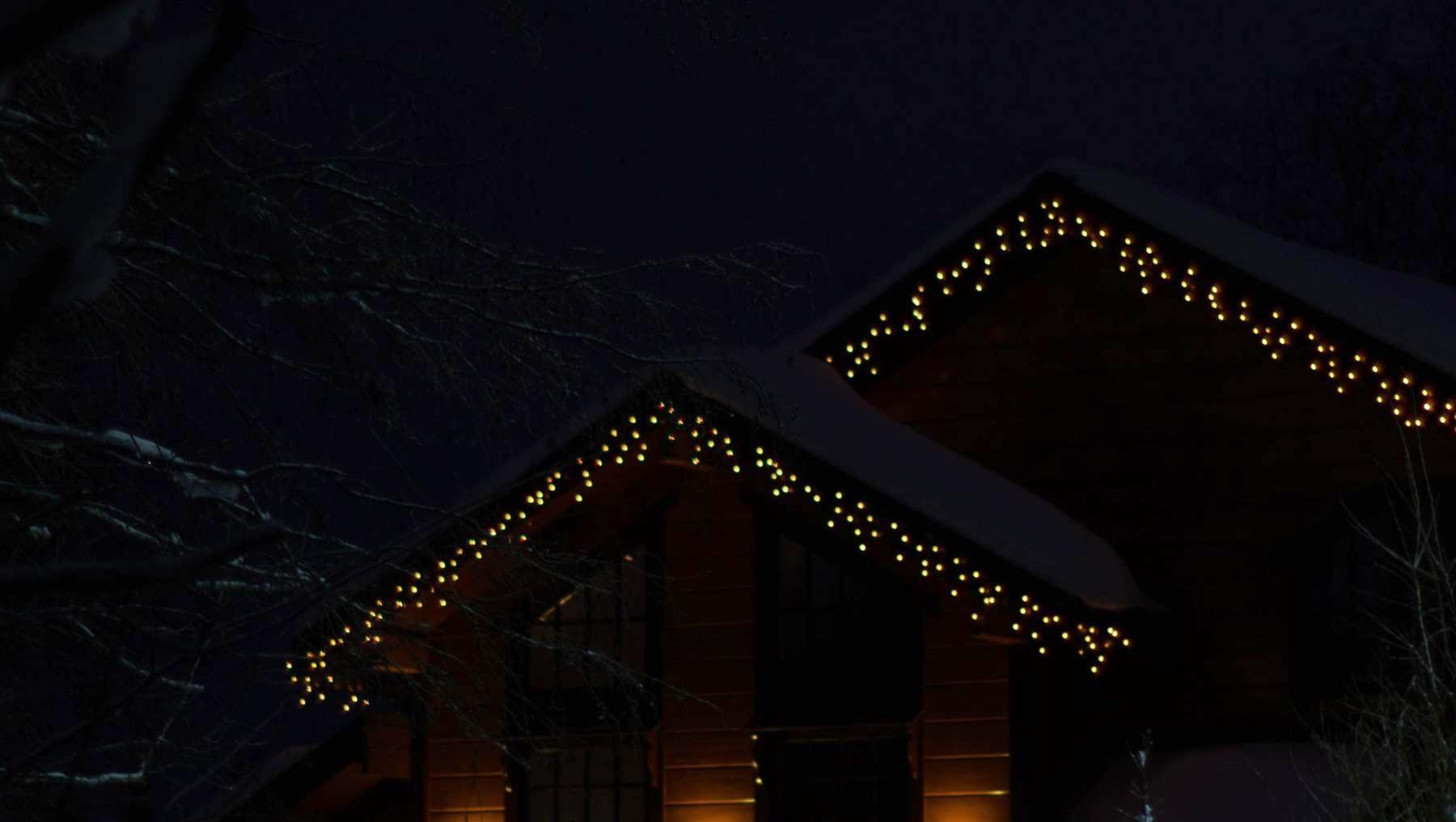 A timber mountain home at night with warm string lights running along both roof gables, surrounded by snow and evergreens.